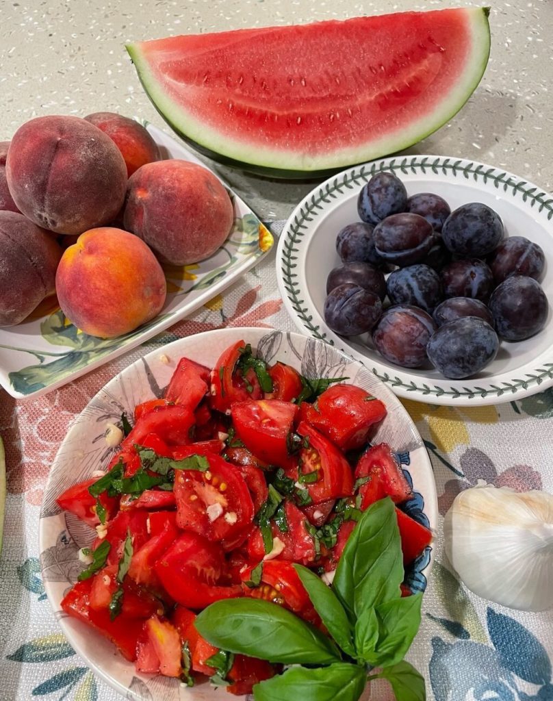 A variety of fresh fruits and vegetables, including watermelon, peaches, plums, and a tomato salad