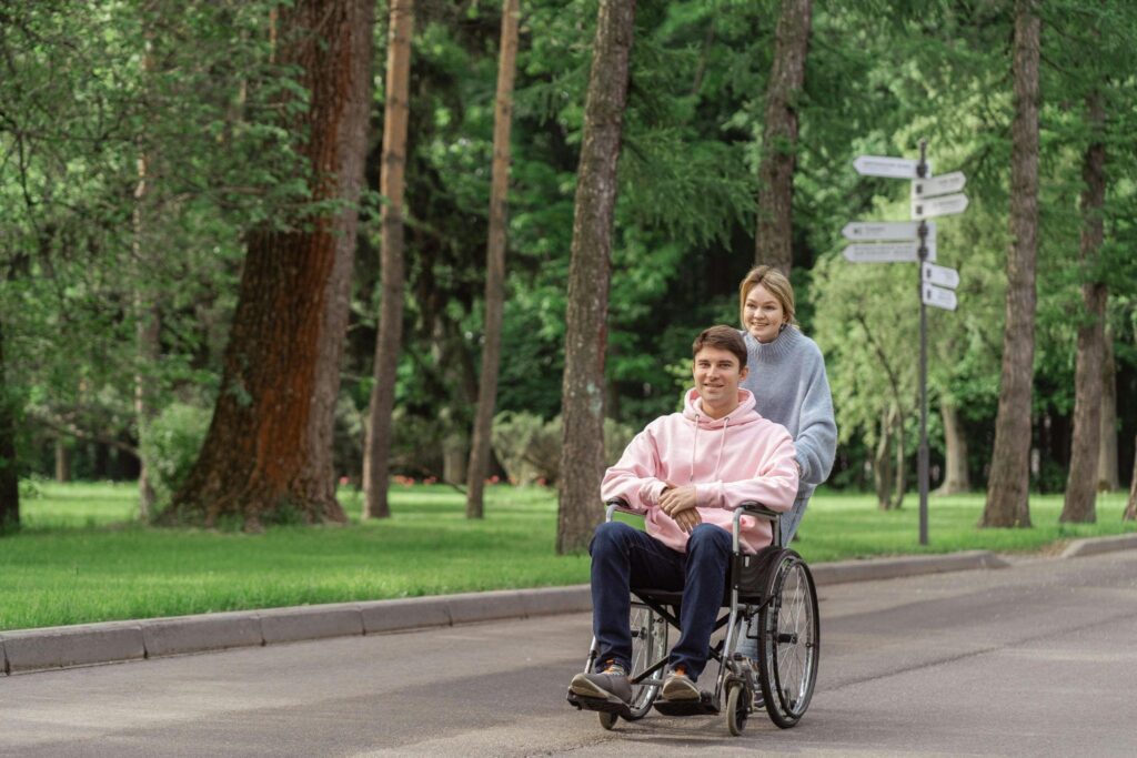 A man in a wheelchair with a woman guiding him at the back, symbolizing support in caring for a disabled spouse.