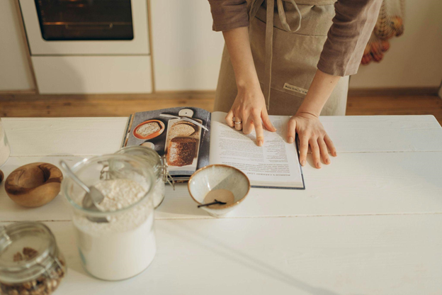 A person in an apron points at a recipe in an open cookbook on a wooden table.