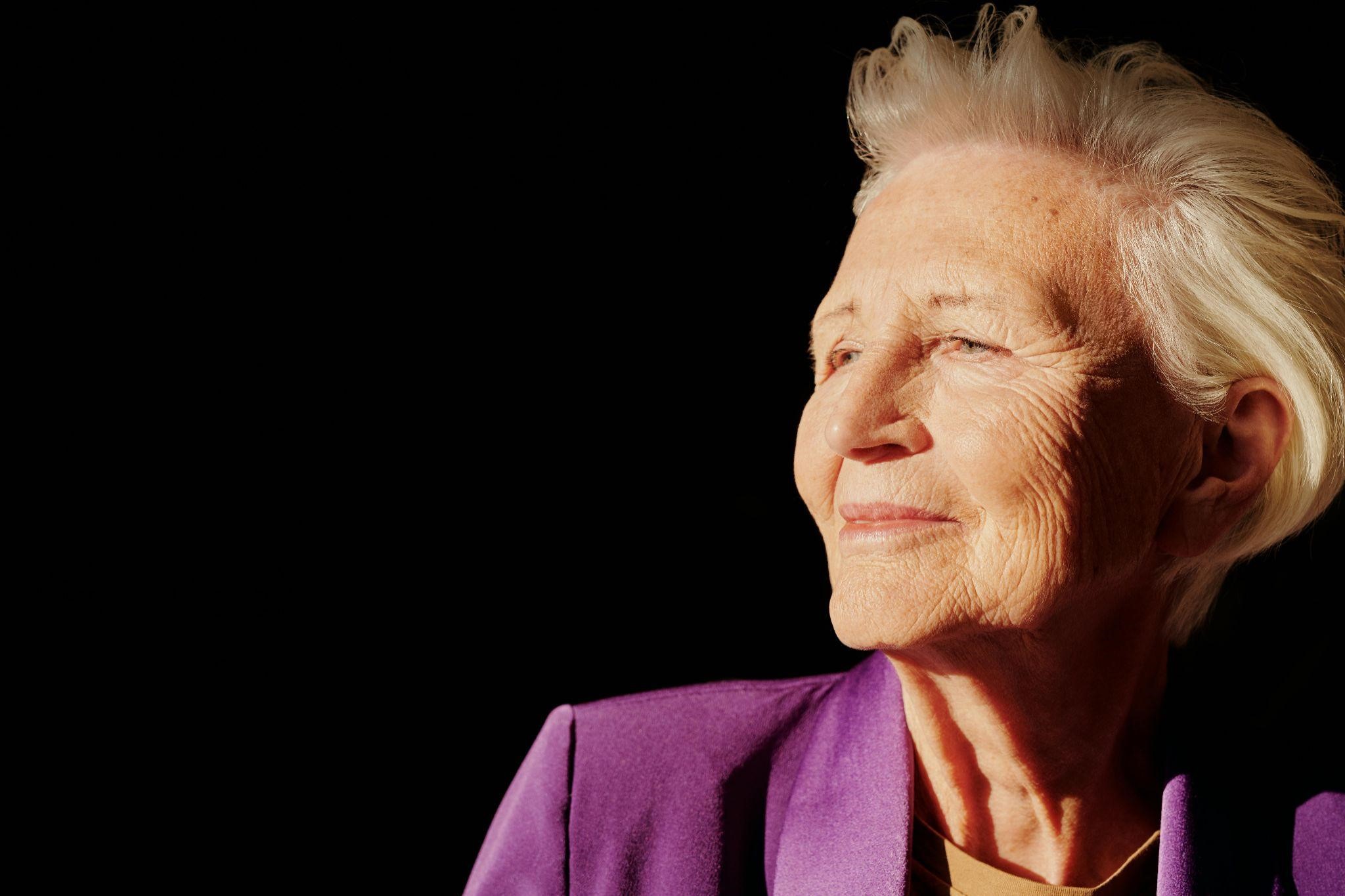 Elderly woman in a bright purple blazer, gazes thoughtfully, exuding the color of alzheimer’s awareness.