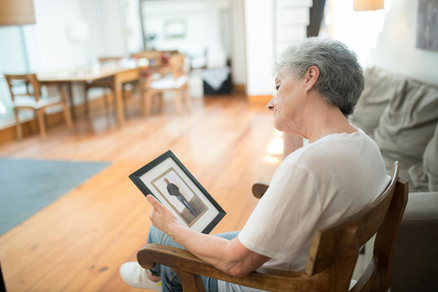 An elderly person sits in a chair, thoughtfully gazing at a framed photograph.