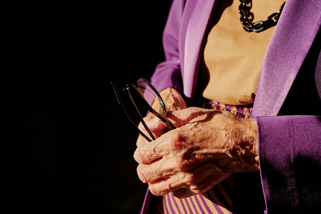 Elderly hands holding glasses, wearing a vibrant purple blazer.