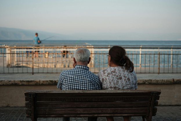 An elderly man and a woman sit side by side on a wooden bench overlooking a calm sea.