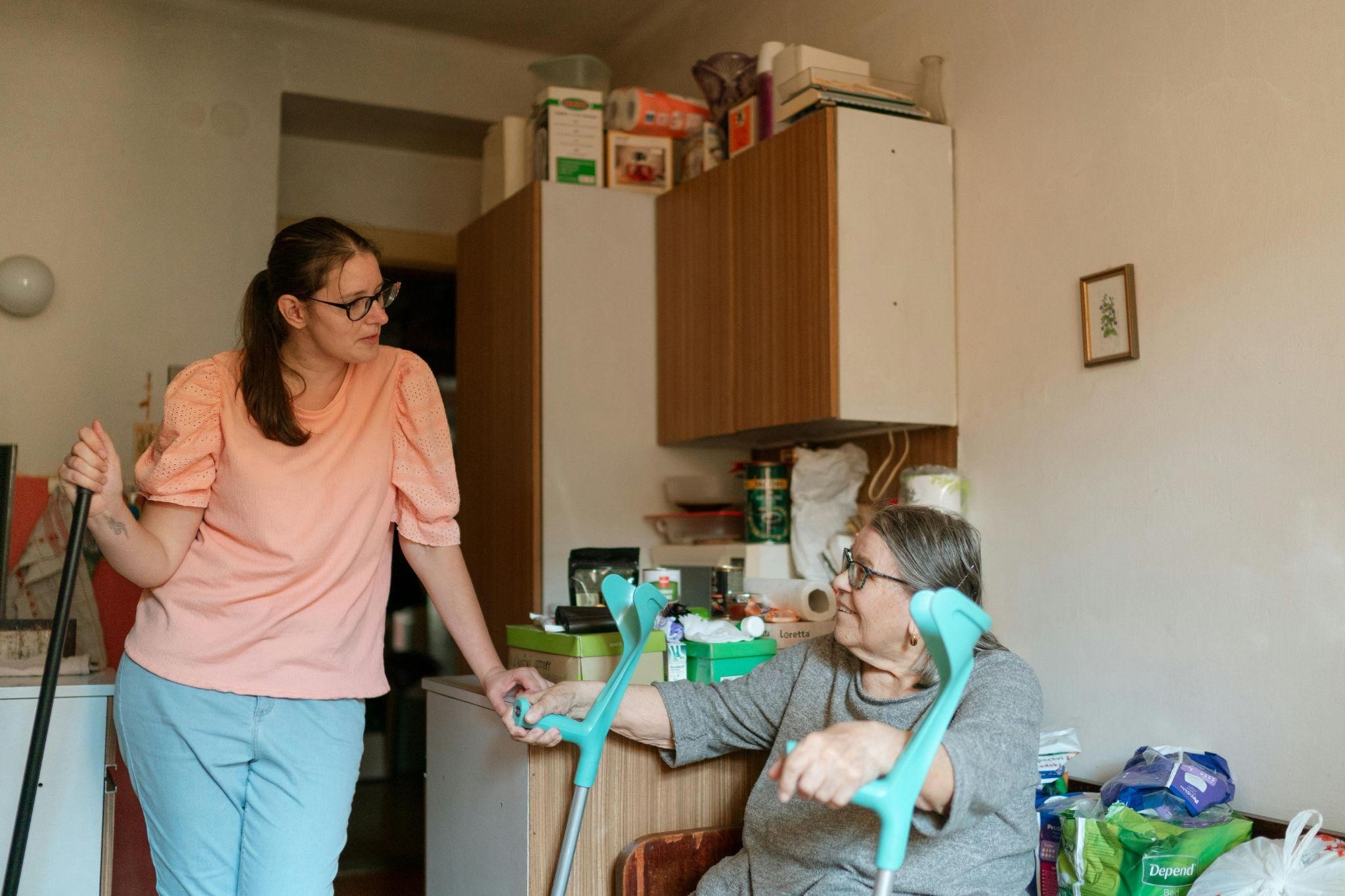 A caregiver leans on a cane, talking to an elderly woman with crutches, conveying why caregiving is hard.