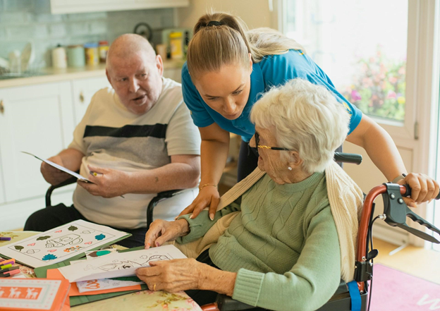 An elderly in a wheelchair receives help from the woman, representing the core role of both caregivers and home health aides.