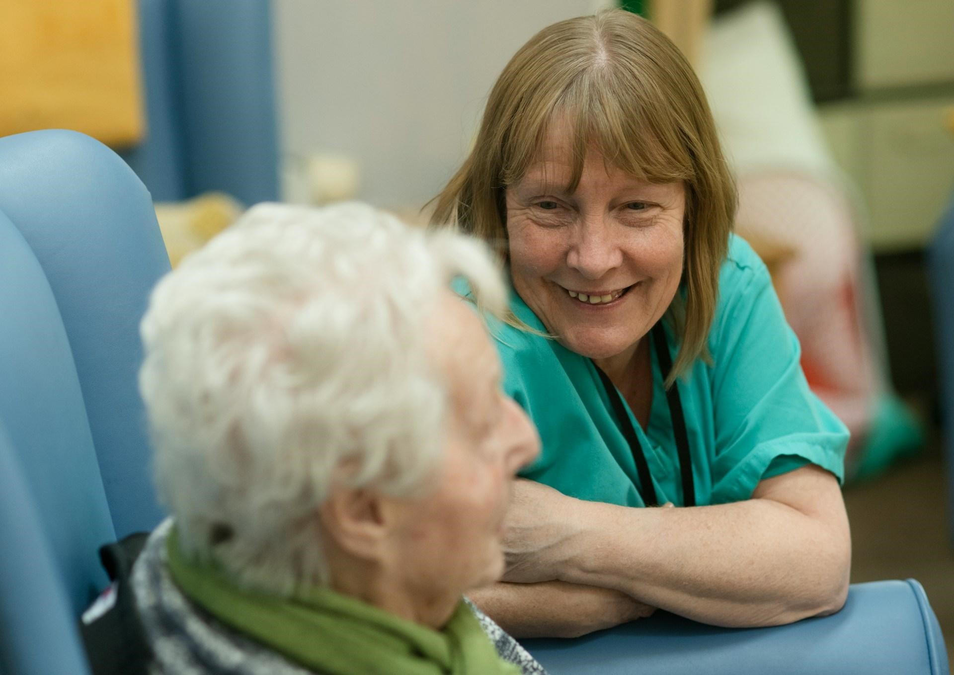 A woman interacts with an older woman seated, emphasizing the supportive environment of standard adult daycare programs.